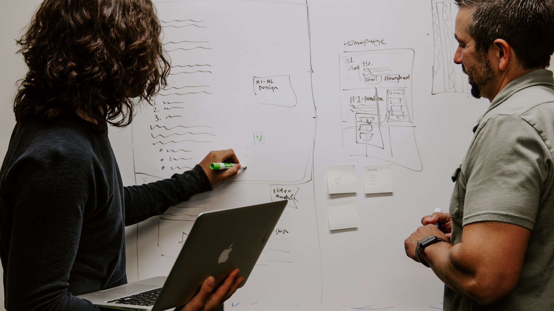 A woman writes something on a whiteboard, a man stands next to her and looks at the whiteboard.